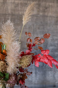 Load image into Gallery viewer, A dried floral arrangement featuring fluffy beige pampas grass, bright red autumn leaves, and muted brown and beige botanicals against a textured gray concrete background.