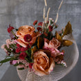 Load image into Gallery viewer, A dried floral arrangement featuring peach and pink roses, deep red chrysanthemums, and beige grasses, wrapped in a textured white raffia hat, set against a gray concrete background.