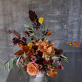 Load image into Gallery viewer, An autumnal floral arrangement in a white cylindrical vase, featuring peach roses, orange ranunculus, dark burgundy leaves, and small yellow and red flowers against a textured gray concrete wall.
