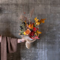 Load image into Gallery viewer, A hand holding a dried flower bouquet with autumnal colors, including orange, red, yellow, and brown elements, wrapped in burlap and paper, set against a textured gray concrete wall.