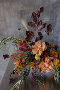 Load image into Gallery viewer, An autumnal floral arrangement featuring peach roses, dark burgundy leaves, red berries, yellow flowers, and green foliage, set against a textured gray concrete wall.
