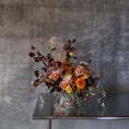 Load image into Gallery viewer, Boise flower delivery. An autumnal floral arrangement in a clear glass vase, featuring orange, peach, yellow, and burgundy flowers with dried pampas grass and dark leaves, displayed on a reflective metal table against a textured gray concrete wall.
