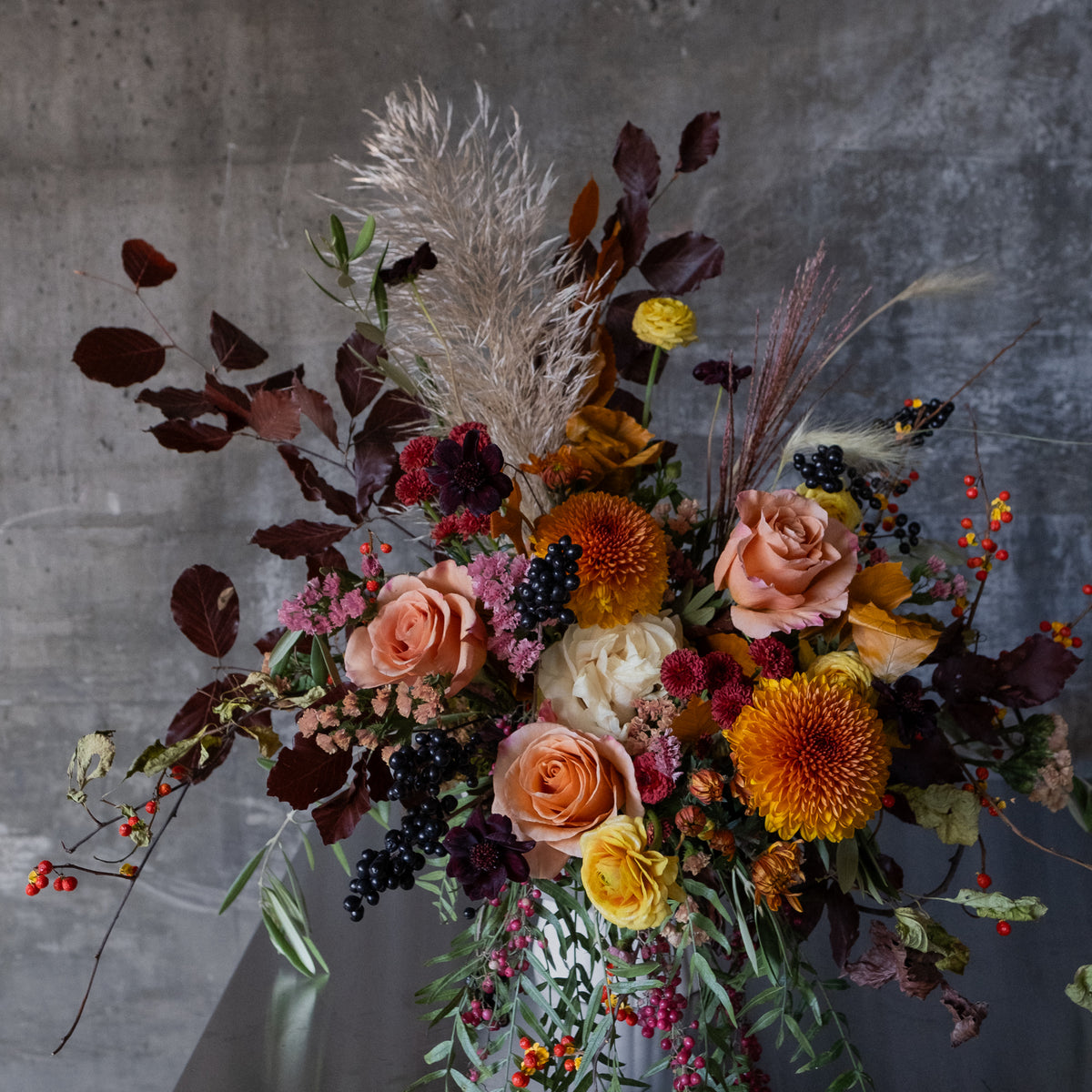A rustic dried flower arrangement in a clear glass vase, featuring peach roses, orange chrysanthemums, pampas grass, and dark burgundy foliage, set against a textured gray concrete wall.