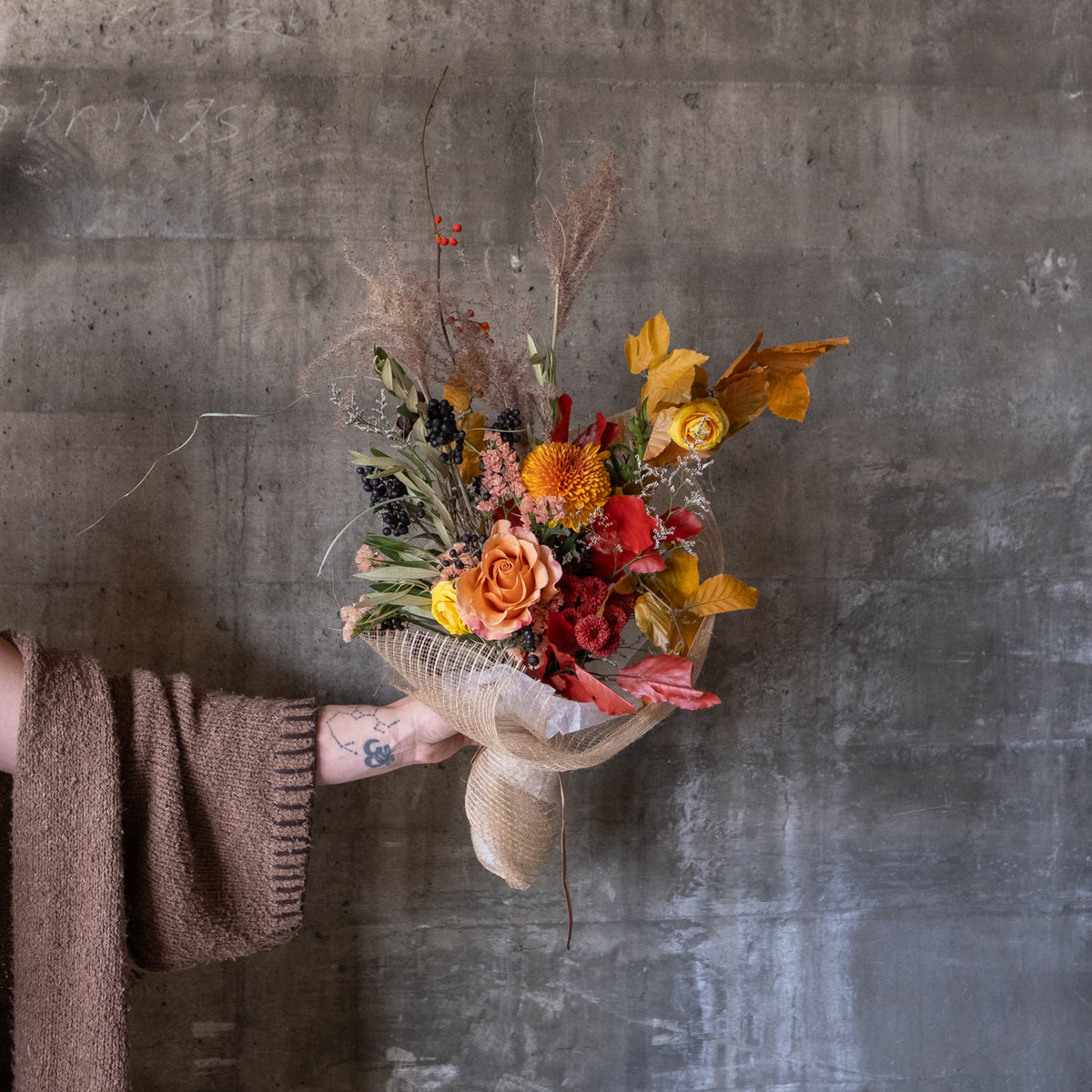 A hand holding a dried flower bouquet with autumnal colors, including orange, red, yellow, and brown elements, wrapped in burlap and paper, set against a textured gray concrete wall.