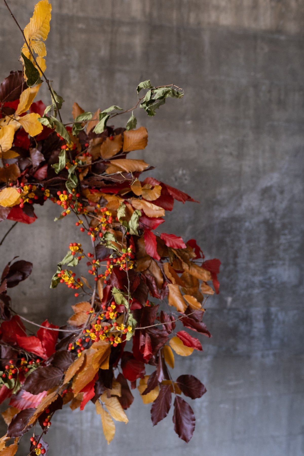 An autumn leaf wreath with red, brown, and yellow foliage and small berries, hanging against a textured gray wall.